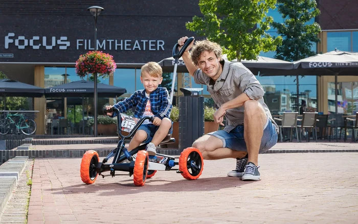 Child on a black pedal kart with chunky orange wheels and blue springs, next to a man holding the push bar outdoors.