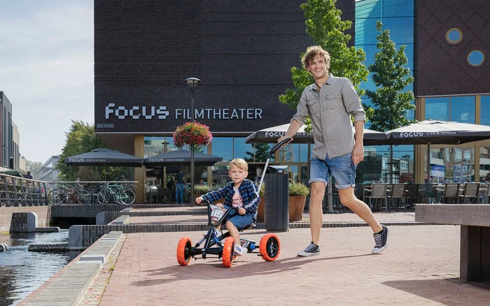 Man pushes a child on a black pedal go-kart with four orange tires and a silver push bar along a brick path.