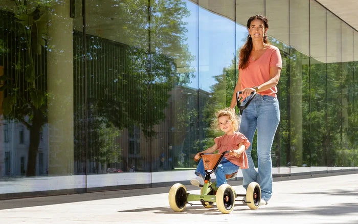 Woman pushing a girl on a green 4-wheel pedal kart with beige tires, brown basket, and a black push handle.