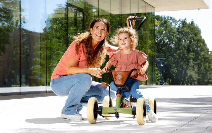 Woman and child with a green retro pedal kart featuring cream wheels, a brown basket, and a push handle.