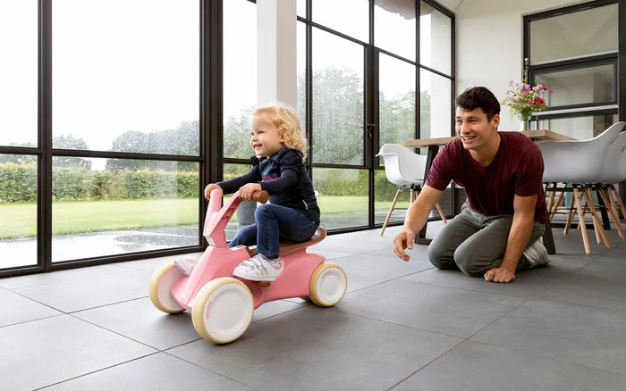 Toddler on a pink retro-style ride-on toy with cream wheels, playing with a man in a modern, sunlit room.