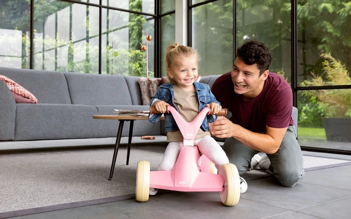 A girl riding a pink four-wheeled toy with cream wheels while a man kneels beside her in a sunlit living room.