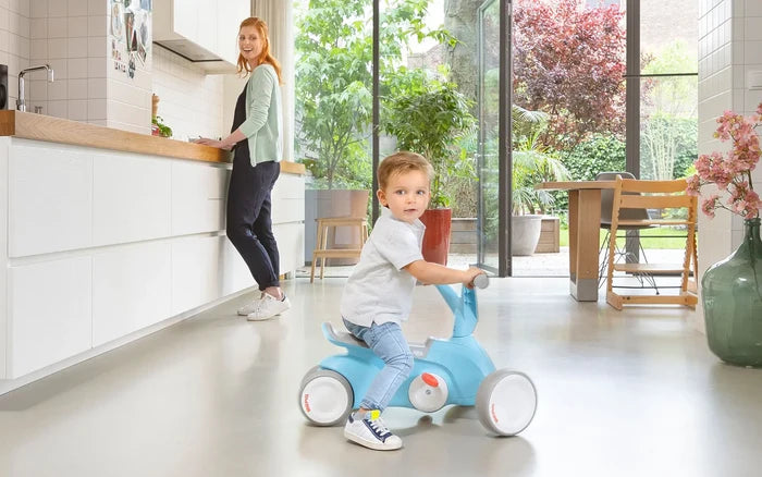 Toddler riding a light blue four-wheel ride-on toy with white wheels in a modern kitchen, woman in background.