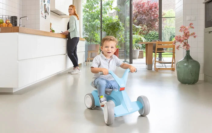 Toddler riding a light blue 4-wheel ride-on toy with grey wheels and blue handlebars in a bright, modern home setting.