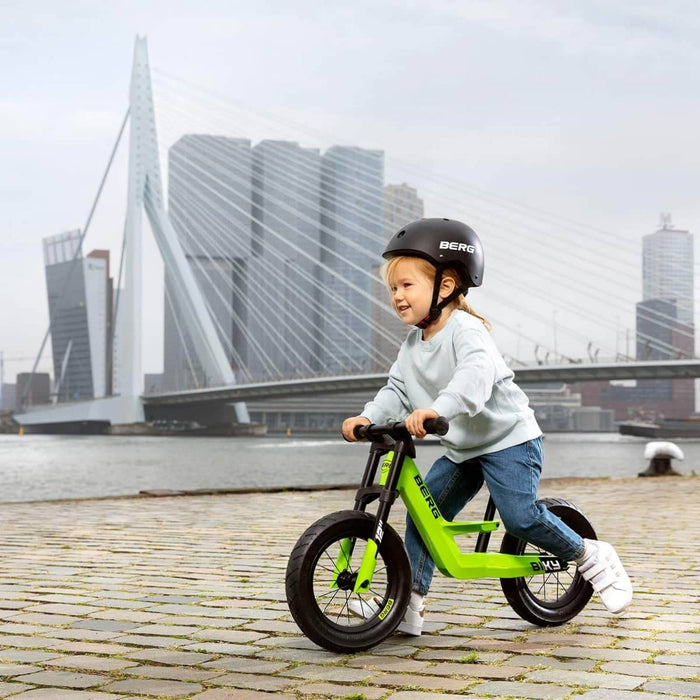 Matte black rounded child's safety helmet with white text and adjustable chin strap, worn by a child on a green bike.