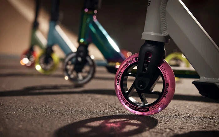 Close-up of a pink translucent scooter wheel with iridescent blue and lime stunt scooters blurred in the background.