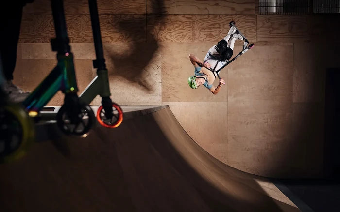 A rider performs a flip in a wooden skatepark, with an iridescent blue and lime stunt scooter in the foreground.