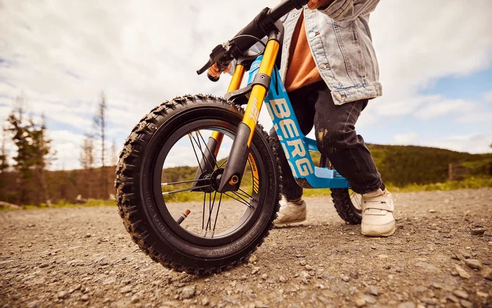 Child on a blue balance bike with gold suspension forks and chunky tires on a gravel path.