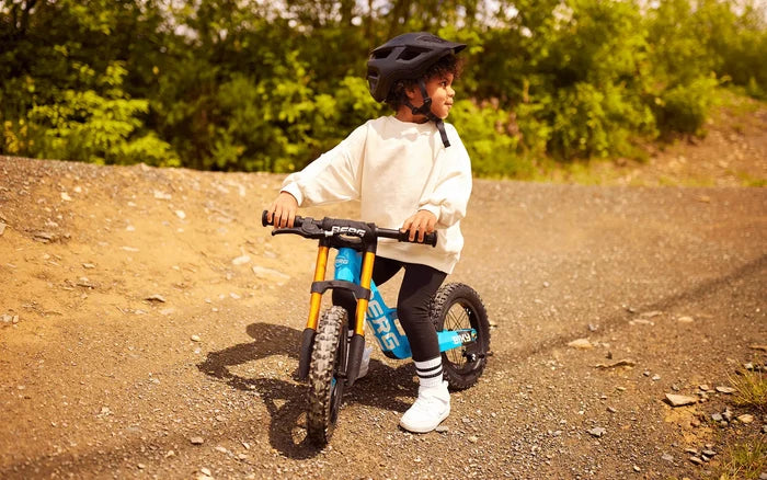Child riding a blue balance bike with gold forks and knobby tires on a gravel path.