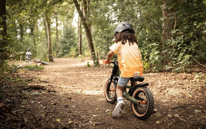 Child riding a green off-road balance bike with orange accents and knobby tires on a wooded trail.