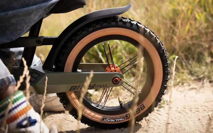 Close-up of olive green balance bike rear wheel with knobby tires, tan sidewalls, and orange hub on a sandy trail.