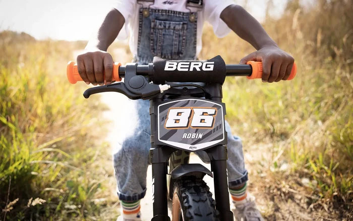 Child on a black balance bike with orange grips, a handbrake, and a racing number plate on a grassy trail.