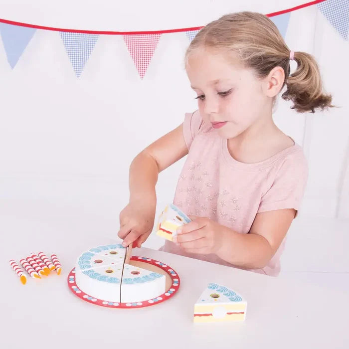 Young girl slicing a wooden toy cake with blue frosting, candles, and a play knife on a red platter.