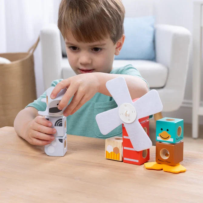 Child stacking colorful wooden blocks with animal faces and a white windmill on a light wood table.