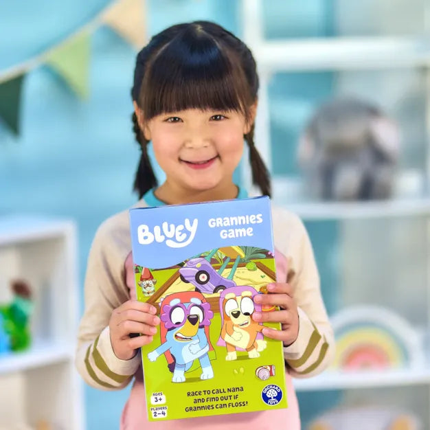 A young girl holds the Bluey Grannies board game box featuring Bluey and Bingo dressed as grandmothers.