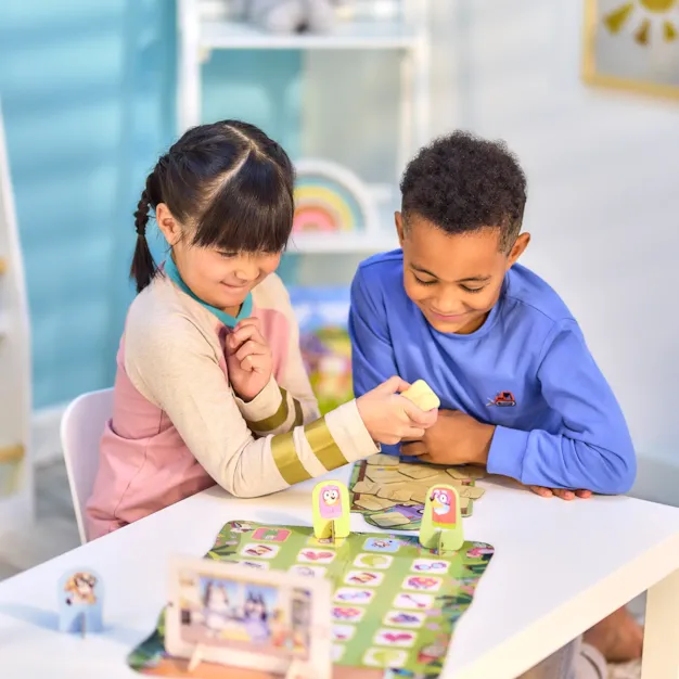 Two children play Bluey Grannies board game with character standees on a colorful green illustrated game board.