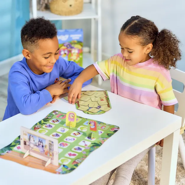 Two children play the Bluey Grannies game with green mats and stand-up character pieces on a white table.