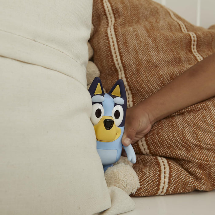 A child's hand holds a Bluey toy figurine peaking out from behind a white pillow and an orange patterned cushion.