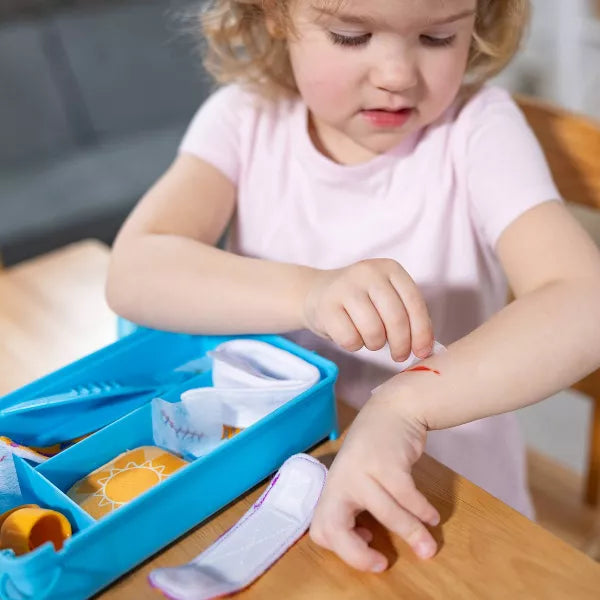 Young girl applies a white fabric bandage to a red mark on her arm from a blue play set tray with cloth medical toys.
