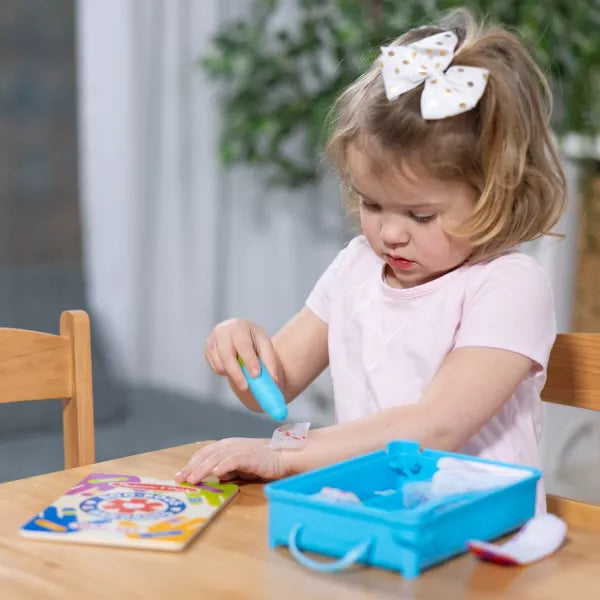 A young girl applies a toy bandage to her arm with a blue applicator from an open blue medical play set.