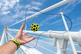 Hand wearing yellow wristband attached by black cord to a yellow Moon Ball with black craters flying in mid-air.