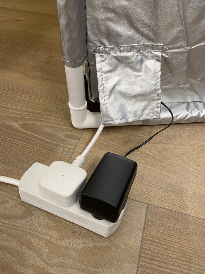 Silver fabric tent corner with white PVC frame and power cords plugged into a power strip on a light wood floor.