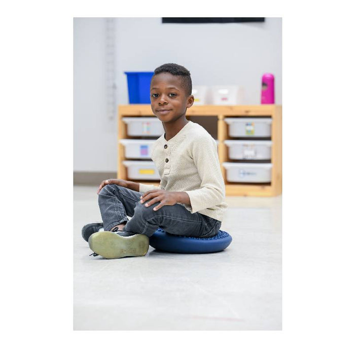 Young boy sitting on a blue, textured, disc-shaped balance cushion on a classroom floor.