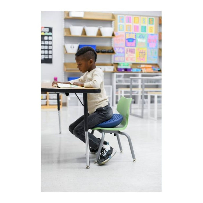 Child sitting on a blue, textured circular cushion on a green chair at a desk in a classroom.