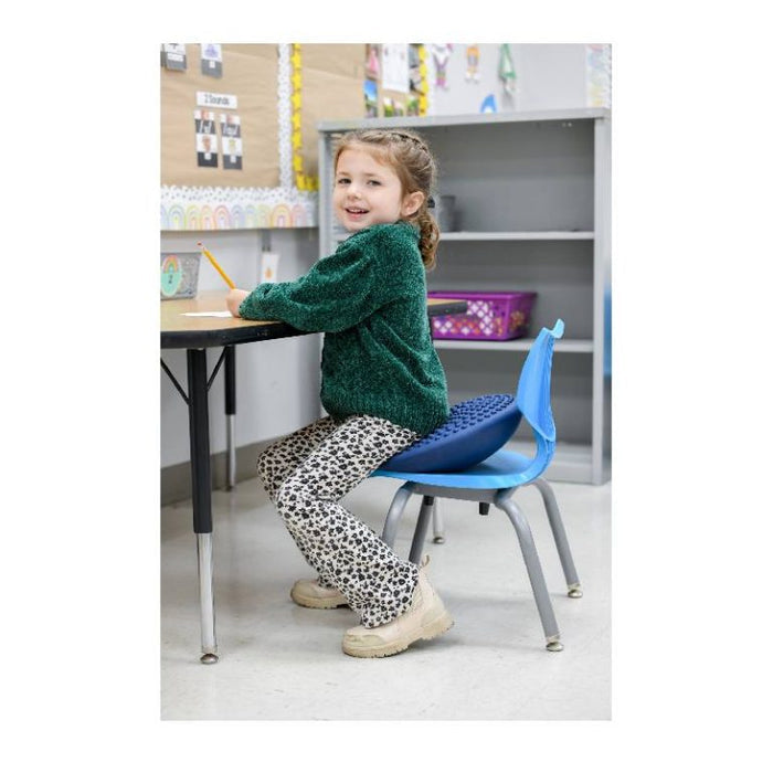 Young girl sitting on a blue round textured cushion on a blue classroom chair while smiling.