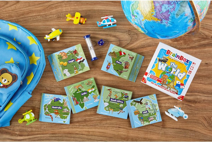 BrainBox The World game box, colorful map cards, globe, and toy planes on a wooden surface near a blue star backpack.