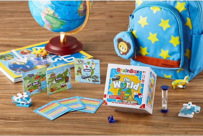 BrainBox The World game box, country cards, and accessories on a wooden table with a globe and star-patterned backpack.