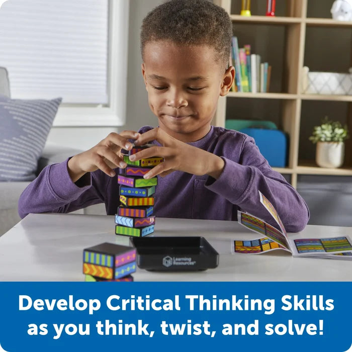 A child builds a tower with colorful, patterned blocks on a white table following an instruction booklet.