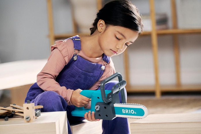 Child uses a chunky teal and grey toy chainsaw with a white blade and BRIO logo to play on wooden blocks.