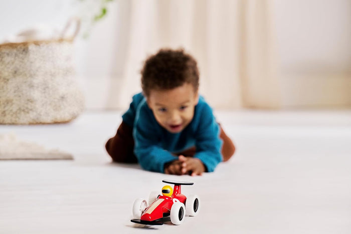 Red toy race car with white wheels and yellow driver, with a blurred child crawling behind it on a white floor.