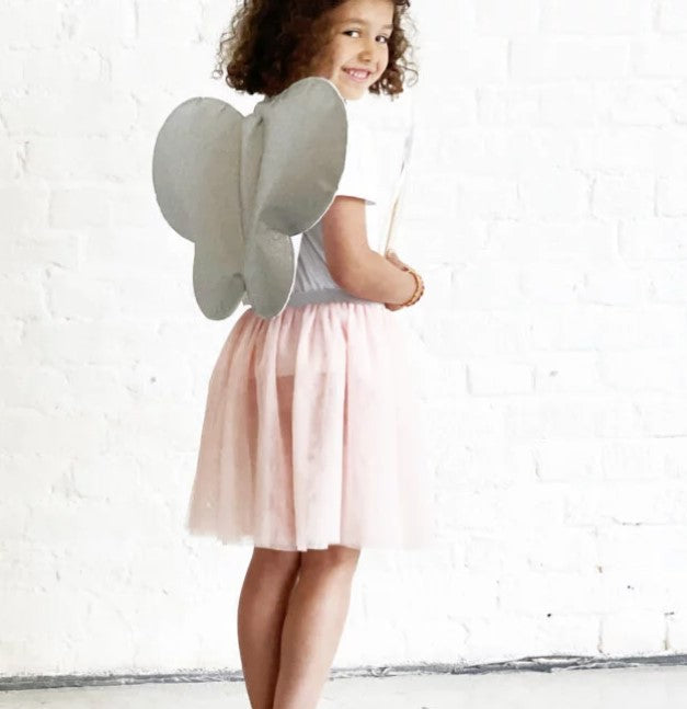 Young girl wearing grey butterfly wings and a light pink tulle tutu skirt against a white brick wall.
