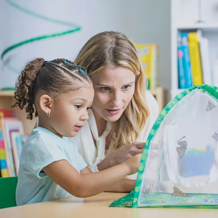 A young girl and woman observe butterflies inside a white mesh cylindrical habitat with green trim.