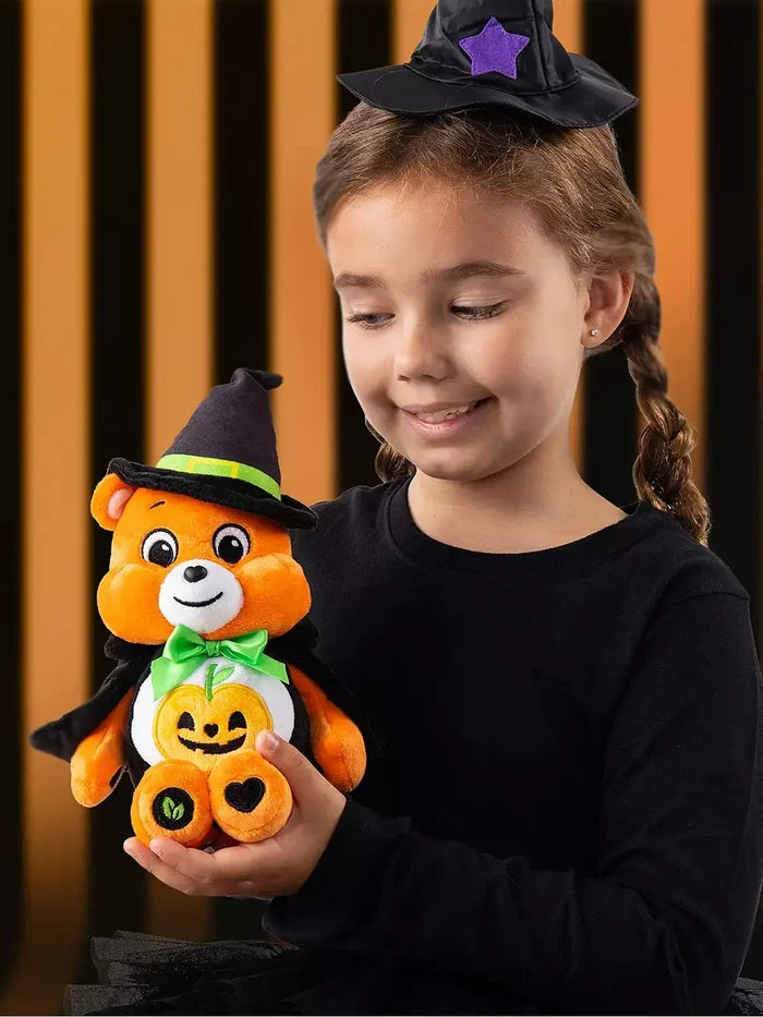 Girl holds an orange Care Bear plush wearing a black witch hat and cape with a pumpkin belly badge.