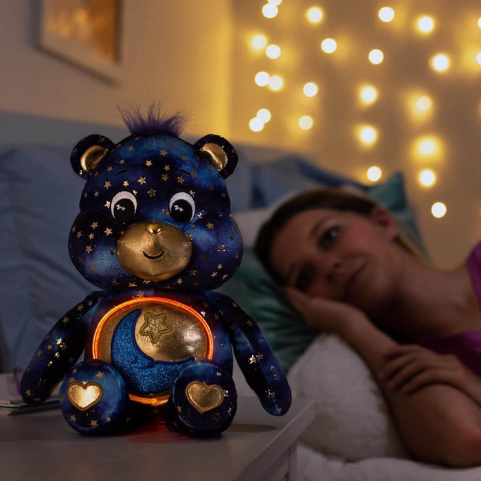 Navy blue galaxy plush bear with gold stars and a glowing moon belly sitting on a bedside table.