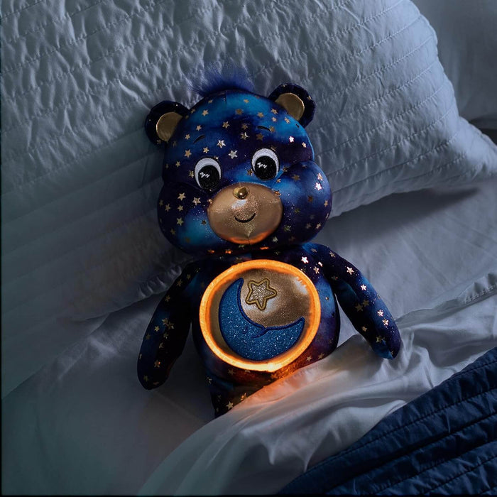 Dark blue starry plush bear with a glowing moon and star belly light, tucked into a white bed with pillows.