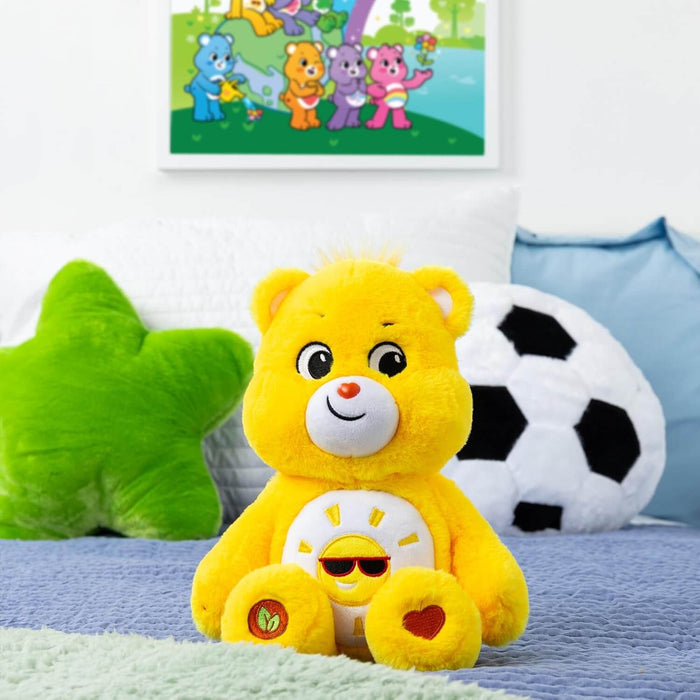 Bright yellow plush bear with a sun belly badge wearing sunglasses, sitting on a bed with star and soccer ball pillows.