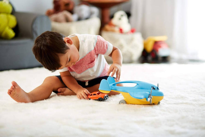 Blue and yellow toy cargo plane with grey propellers and a top handle, shown with a child playing on a white rug.