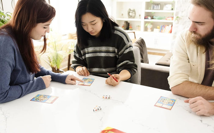 Three people play Catan Dice Game with resource dice and hexagonal map scorecards on a bright white table.