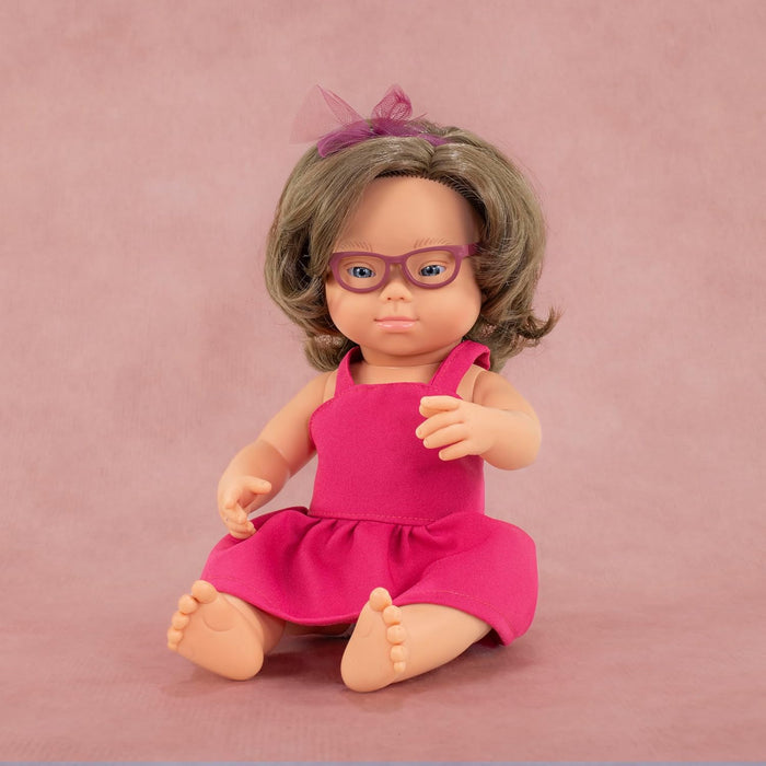 Caucasian doll with Down syndrome features, short brown hair, purple glasses, and a pink dress on a pink background.