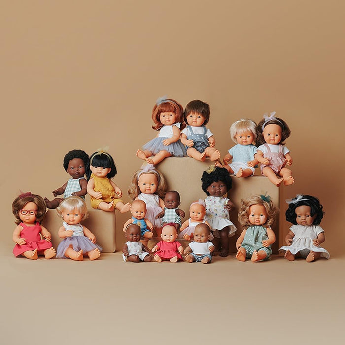 Inclusive baby doll with red glasses and a red dress, pictured with a diverse group of dolls on a tan background.