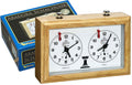 Light wood mechanical chess clock with two white analog faces and red flags, shown with its blue brand box.