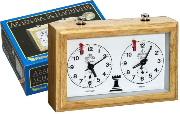 Light wood mechanical chess clock with two white analog faces and red flags, shown with its blue brand box.