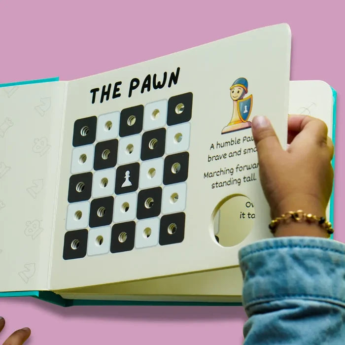 Child turns a page in an open board book titled THE PAWN, featuring a chess grid and a cartoon character illustration.