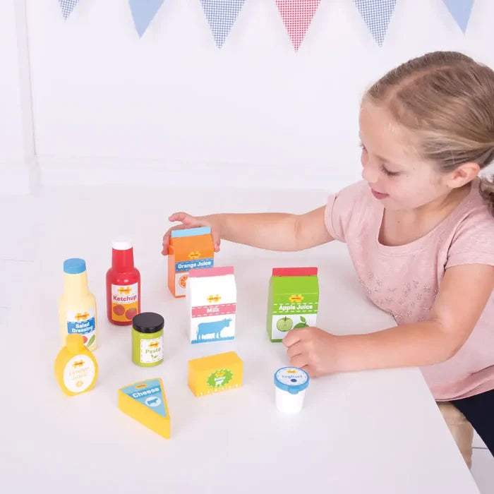 Child playing with wooden toy groceries: milk, juice cartons, cheese wedge, and colorful condiment bottles.