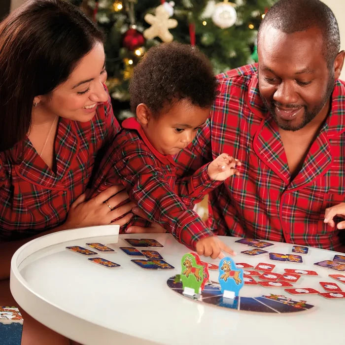 Family in red plaid pajamas plays a reindeer board game with colorful cards on a white table near a Christmas tree.