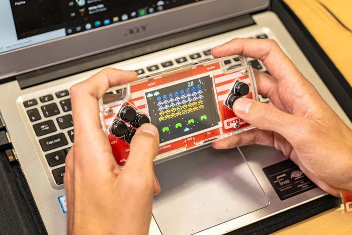 Hands holding a clear handheld console with red stripes and black buttons, showing a pixelated arcade game on screen.
